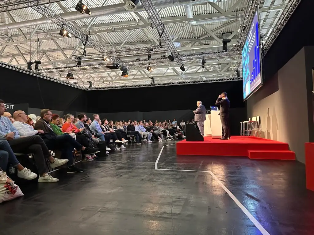In einem modernen Konferenzsaal hält ein Redner auf einer leuchtend roten Bühne einen Vortrag vor einem großen Publikum. Die Teilnehmer sitzen in Stuhlreihen und hören aufmerksam zu. Deckenlampen erhellen den Raum und auf einem großen Bildschirm werden visuelle Elemente der Präsentation angezeigt.
