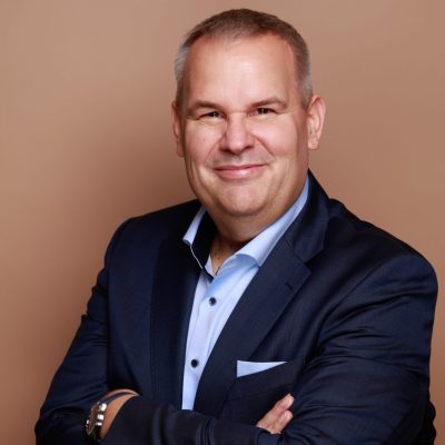 Ein Mann mittleren Alters mit kurzen grauen Haaren, der einen dunkelblauen Anzug und ein hellblaues Hemd trägt, steht mit verschränkten Armen und lächelnd vor einem einfarbig braunen Hintergrund.