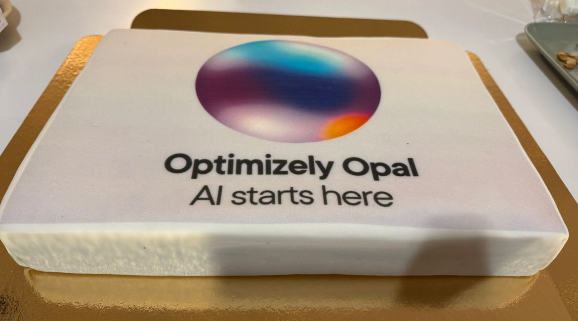 Ein rechteckiger Kuchen mit weißem Zuckerguss, auf dem ein buntes, kreisförmiges Logo und der Text "Optimizely Opal AI starts here" aufgedruckt sind. Die Torte steht auf einem goldenen Brett.